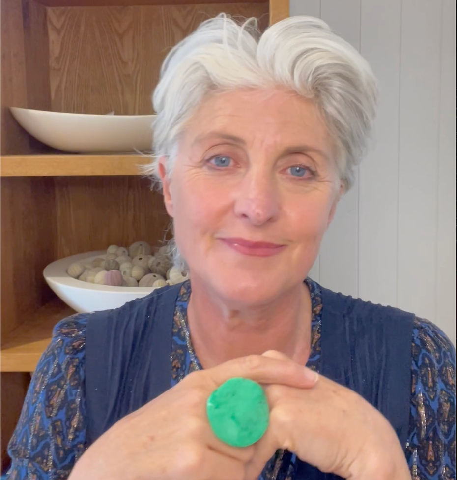 Mature woman with grey hair, blue eyes, green statement ring, smiling indoors with wooden shelves.