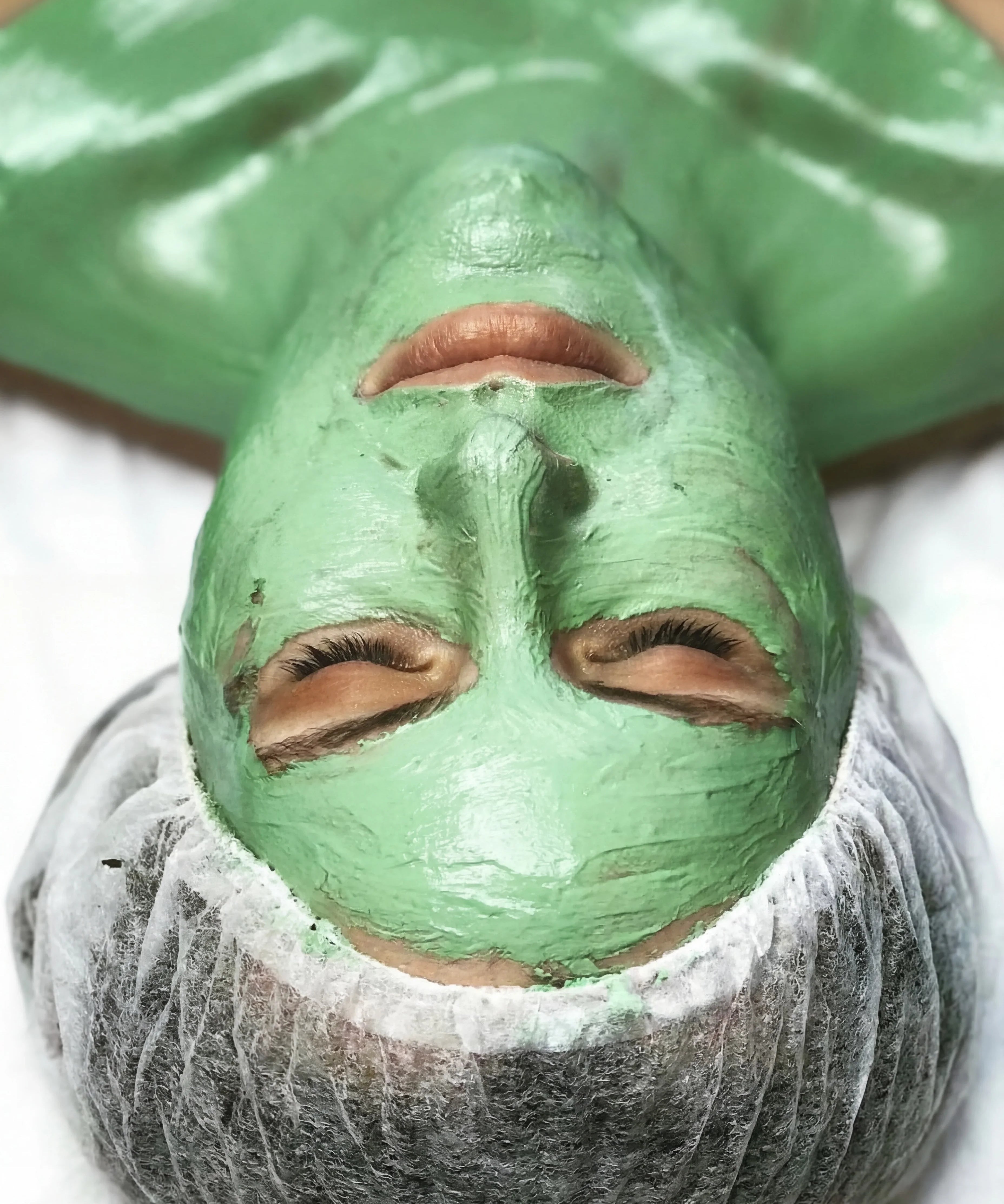 Woman with closed eyes wearing a green clay facial mask and a hair cap, spa skincare treatment