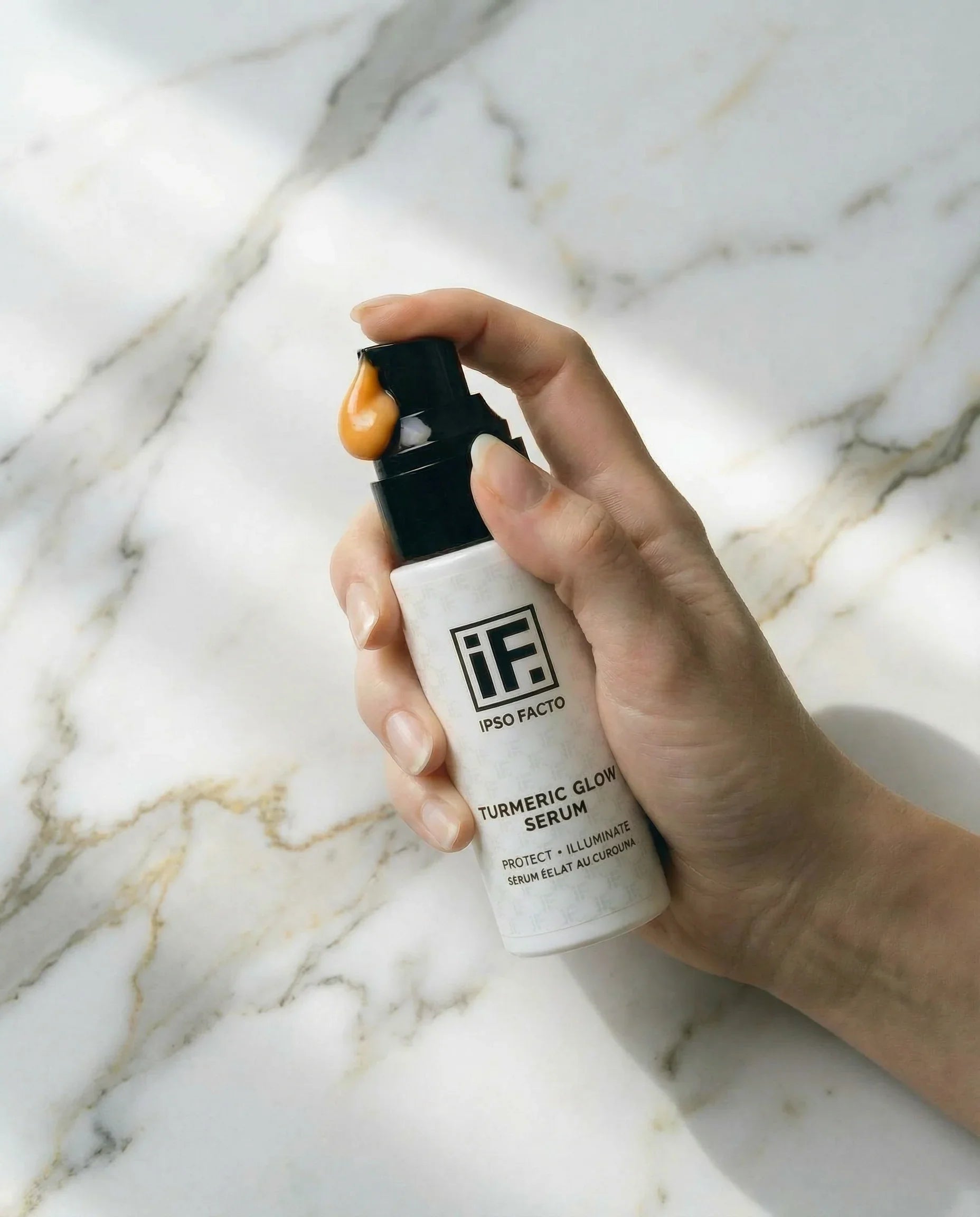 Hand holding IF Turmeric Glow Serum bottle with golden serum on pump, white marble background